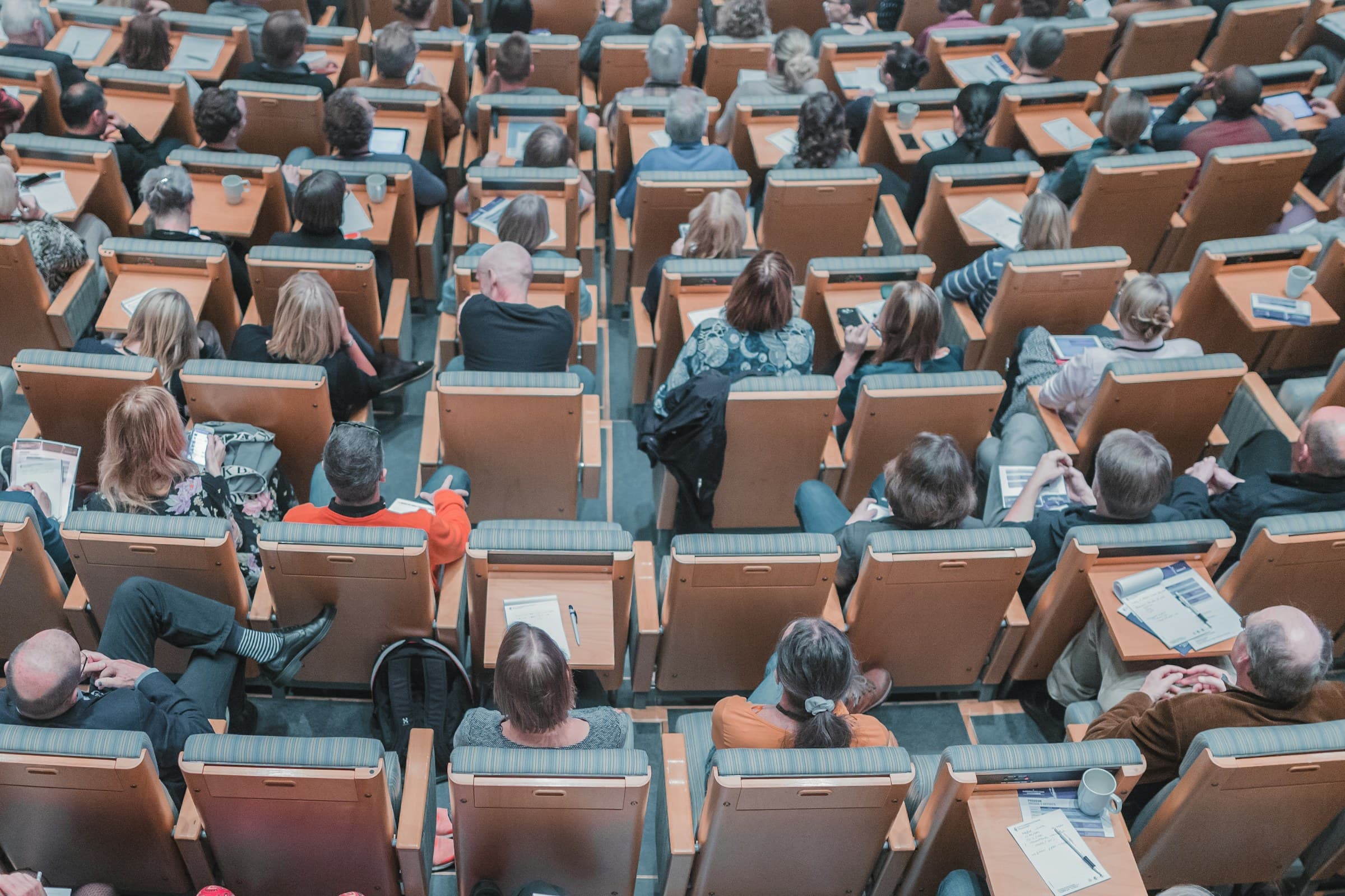 A lecture hall seen from above, rows of seats filled with attendees and papers on the desks.