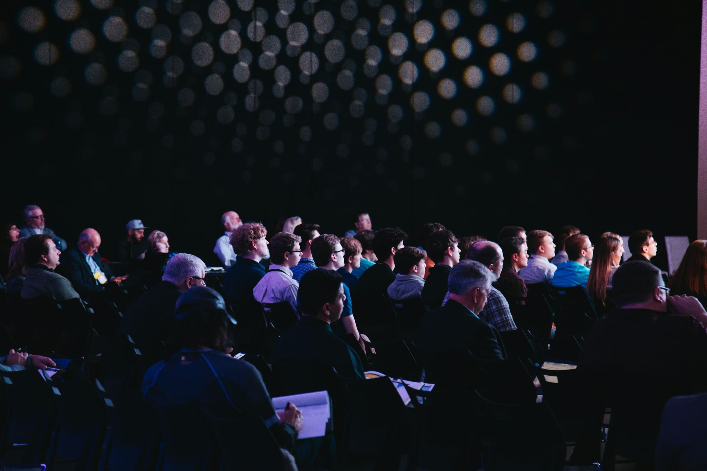 An attentive audience seated in a darkened conference hall, faces lit by the stage.