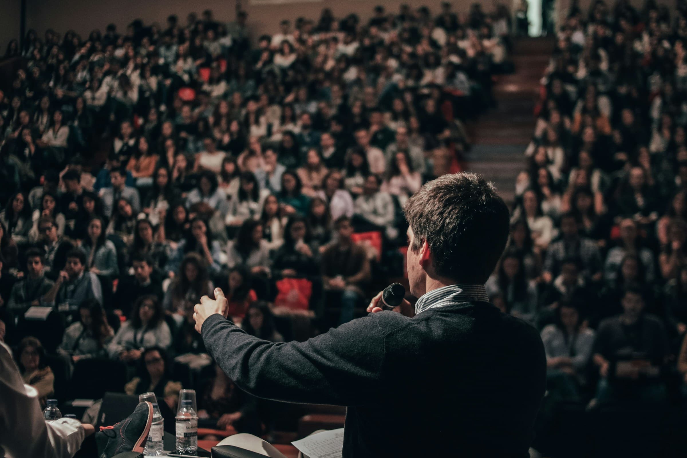A speaker seen from behind, addressing a full auditorium with a microphone in hand.